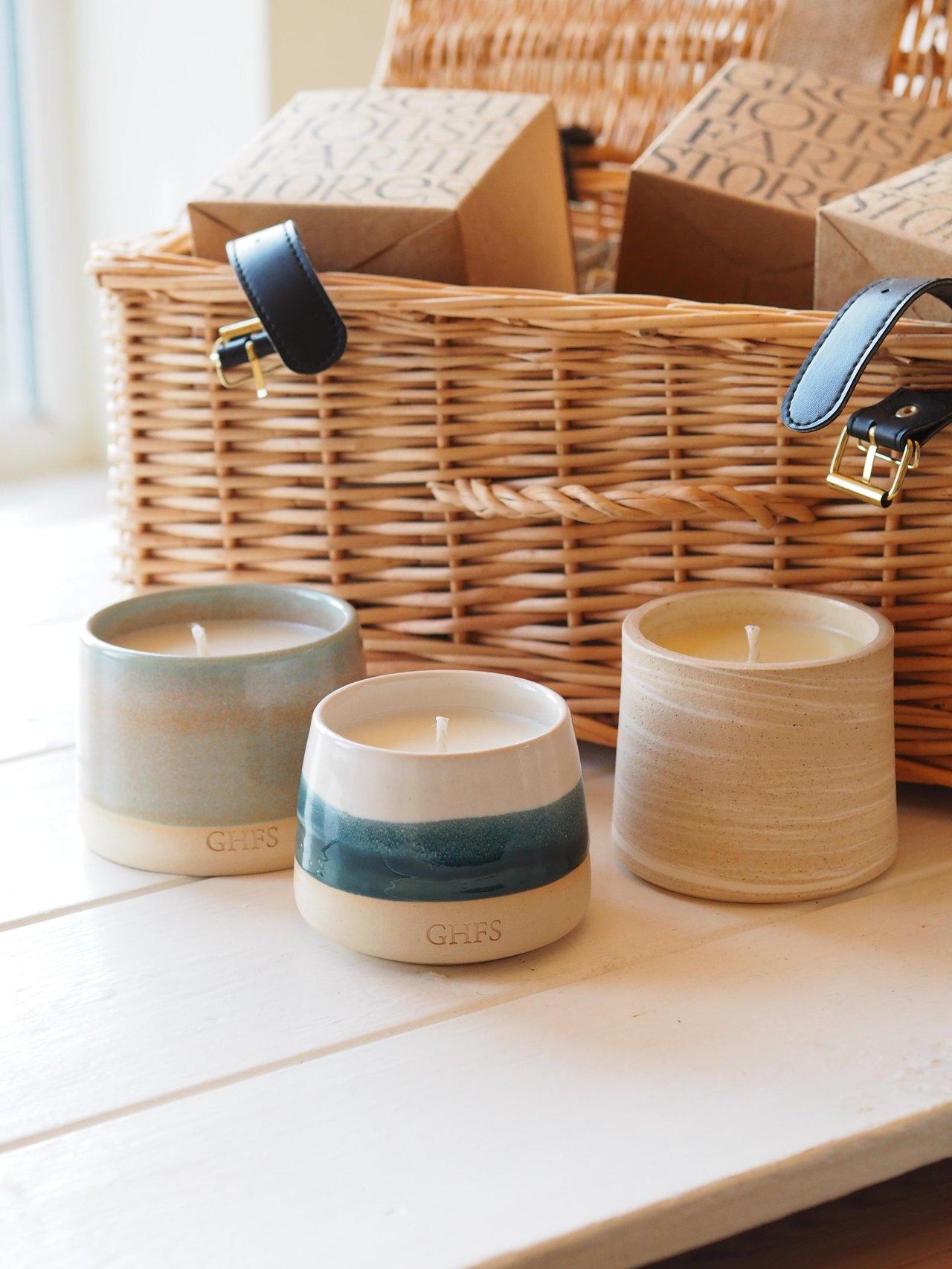 An open willow gift hamper with three luxury candles in clay pots positioned in front of it.