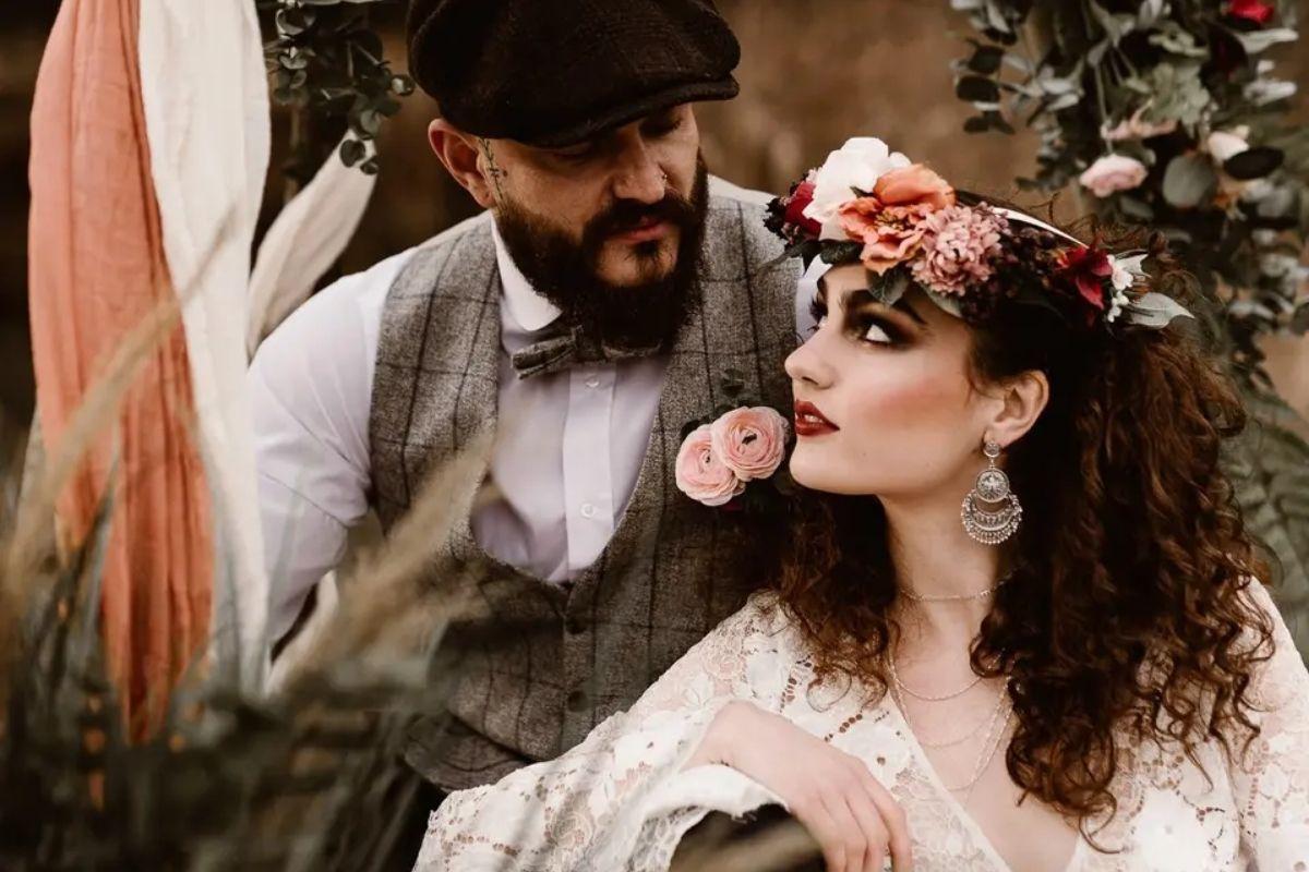a bride with dramatic makeup and a floral crown looks up at her groom who has a tweed suit and beard