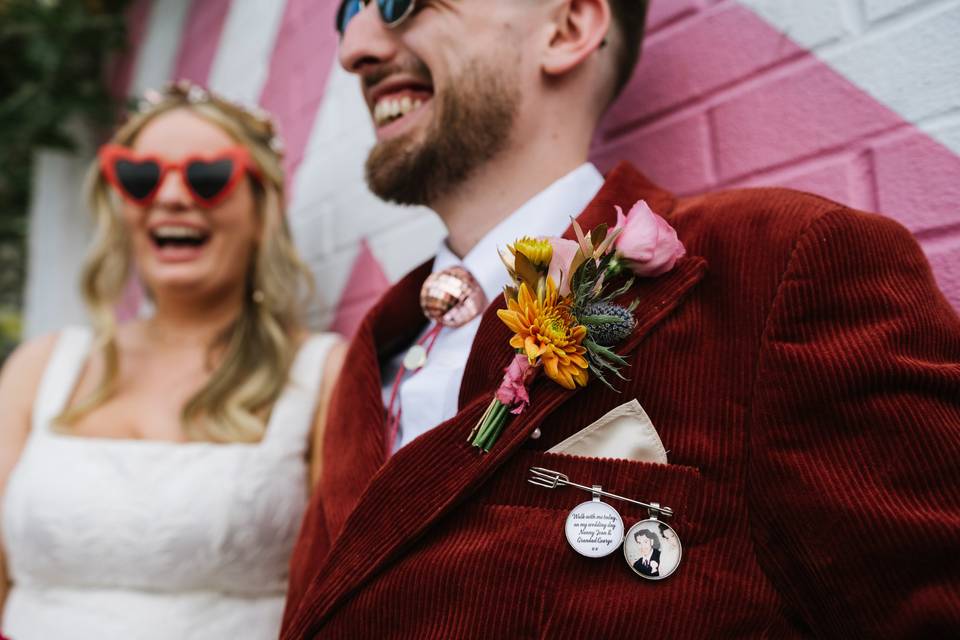 Modern bride with blonde wavy hair wearig a white wedding dress and red heart-shaped sunglasses and a modern groom wearing a white shirt, disco ball bolo tie and a dark red cord suit jacket with a modern buttonhole with yellow and pink flowers and a sentimental photo pin on the suit jacket pocket 