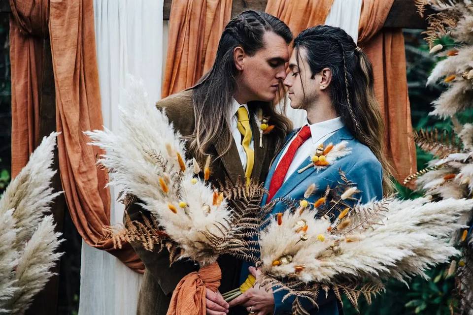 Two grooms, both wearing rustic wedding suits with braided hair gaze into each other's eyes while holding pampas grass bouquets on their wedding day