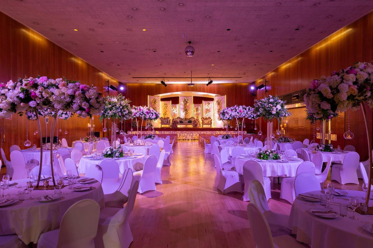 Image of a hall wedding venue set up for an Asian wedding reception complete with tall floral centrepieces and stage set up for the couple.