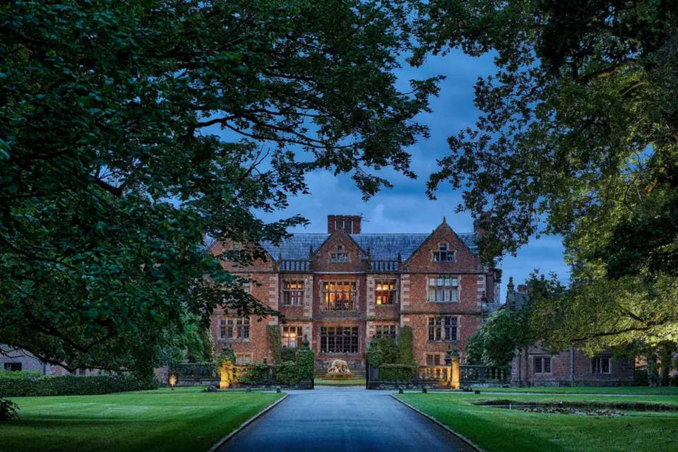 A front view of a grand Cheshire wedding venue with a long drive leading up to it, pictured at dusk with lights pointing at the venue
