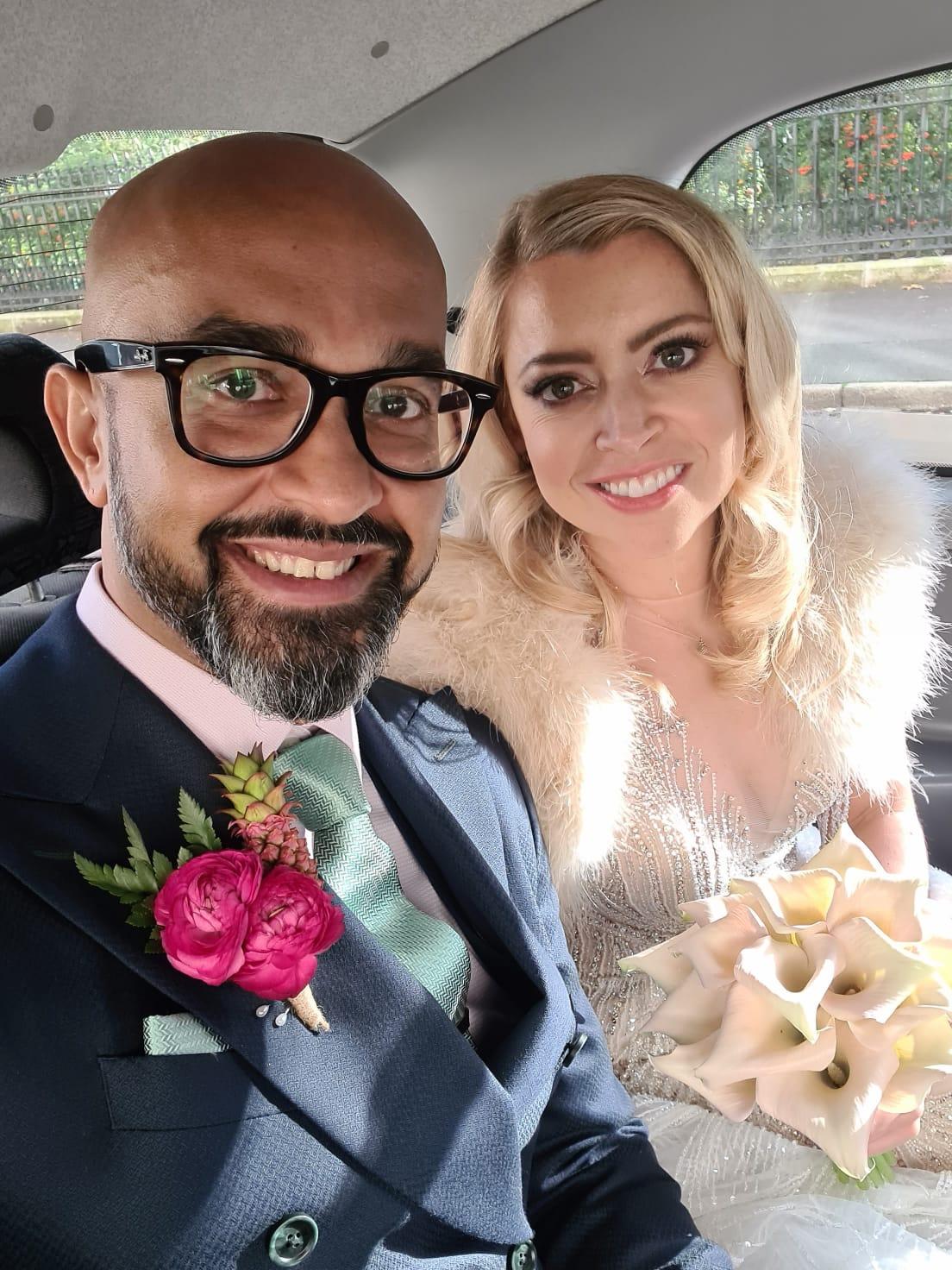 Selfie of a newlywed couple in the back of a wedding car with the brown-skinned groom wearing black rimmed glasses with a light pink shirt, bright pink buttonhole and dark suit and the blonde haired white bride wearing a beaded wedding gown and ivory fur cape