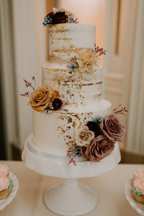 Three tiered white rustic wedding cake with metallic gold leaf and flowers