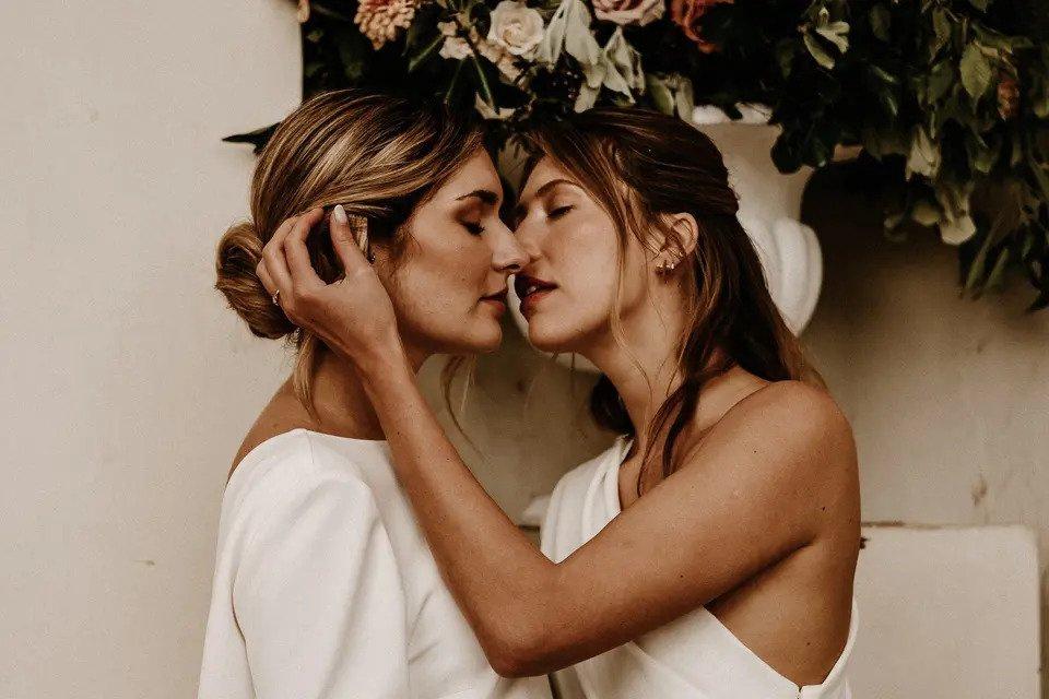 Two brides embrace and lean in for a kiss in front of flowers and a white wall