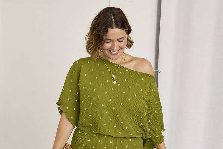 A plus size mother of the bride wearing a green off the shoulder plisse dress with gold polka dots. She is carrying a gold clutch bag and looking downwards while smiling.