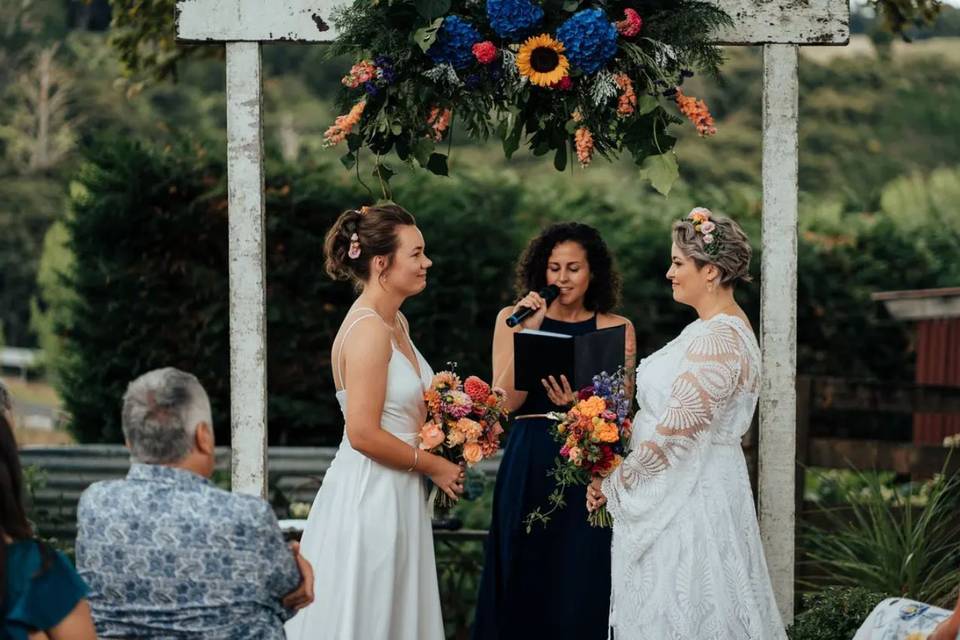 a bridesmaid giving a reading in front of two brides getting married underneath a floral wooden wedding arch