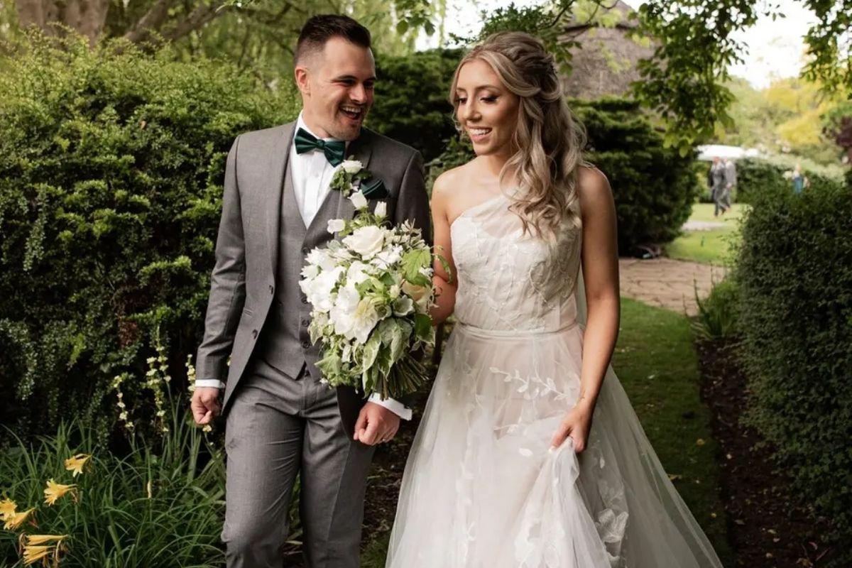 a bride in a beaded wedding dress walks through a wedding venue's gardens with her groom as she holds her white bouquet