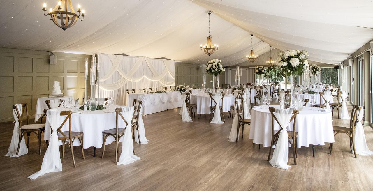 Interior of a marquee-style space set up for a wedding reception with round tables and white drapes.