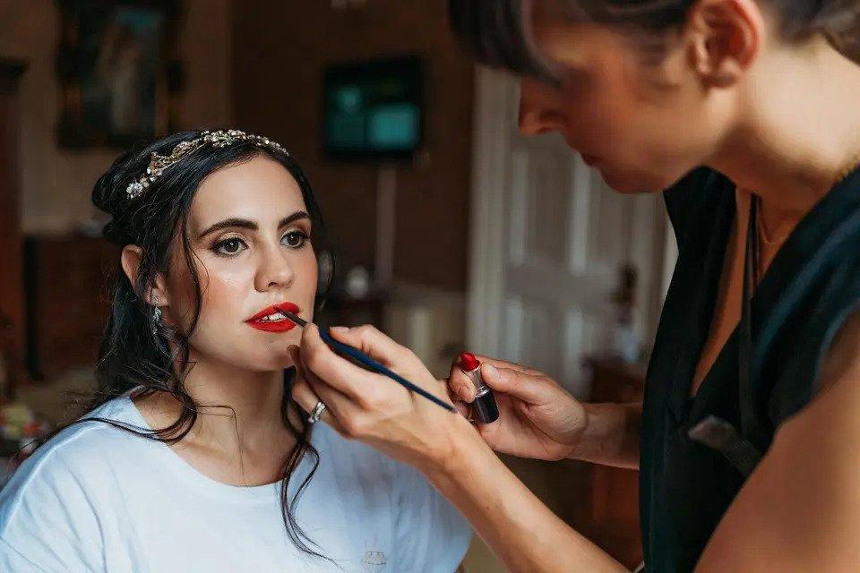 A bride with a headpiece and earrings sits while an MUA applies red lipstick
