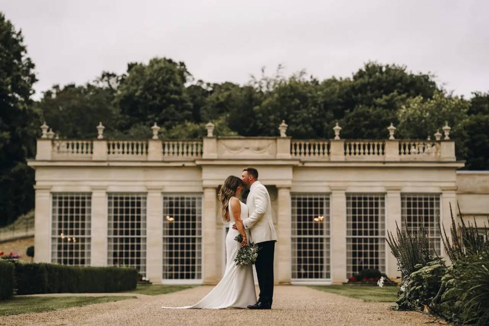 a couple kissing in front of a large orangery wedding venue built from white stone with a gravel pathway leading up to it