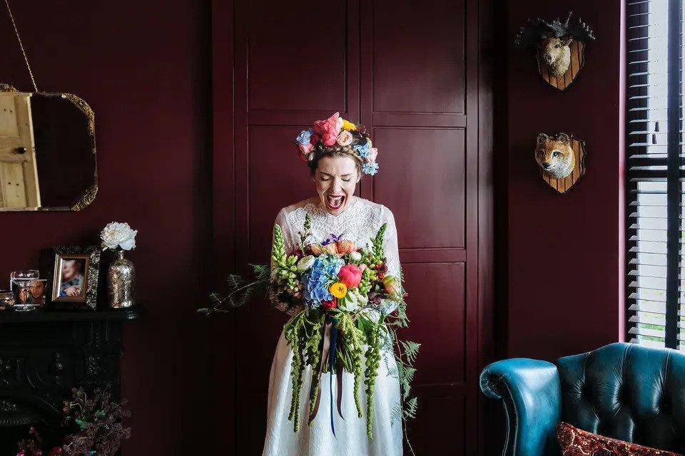A bride standing in front of a wood panelled wall holds a colourful bouquet and wears a colourful flower crown while closing her eyes and opening her mouth