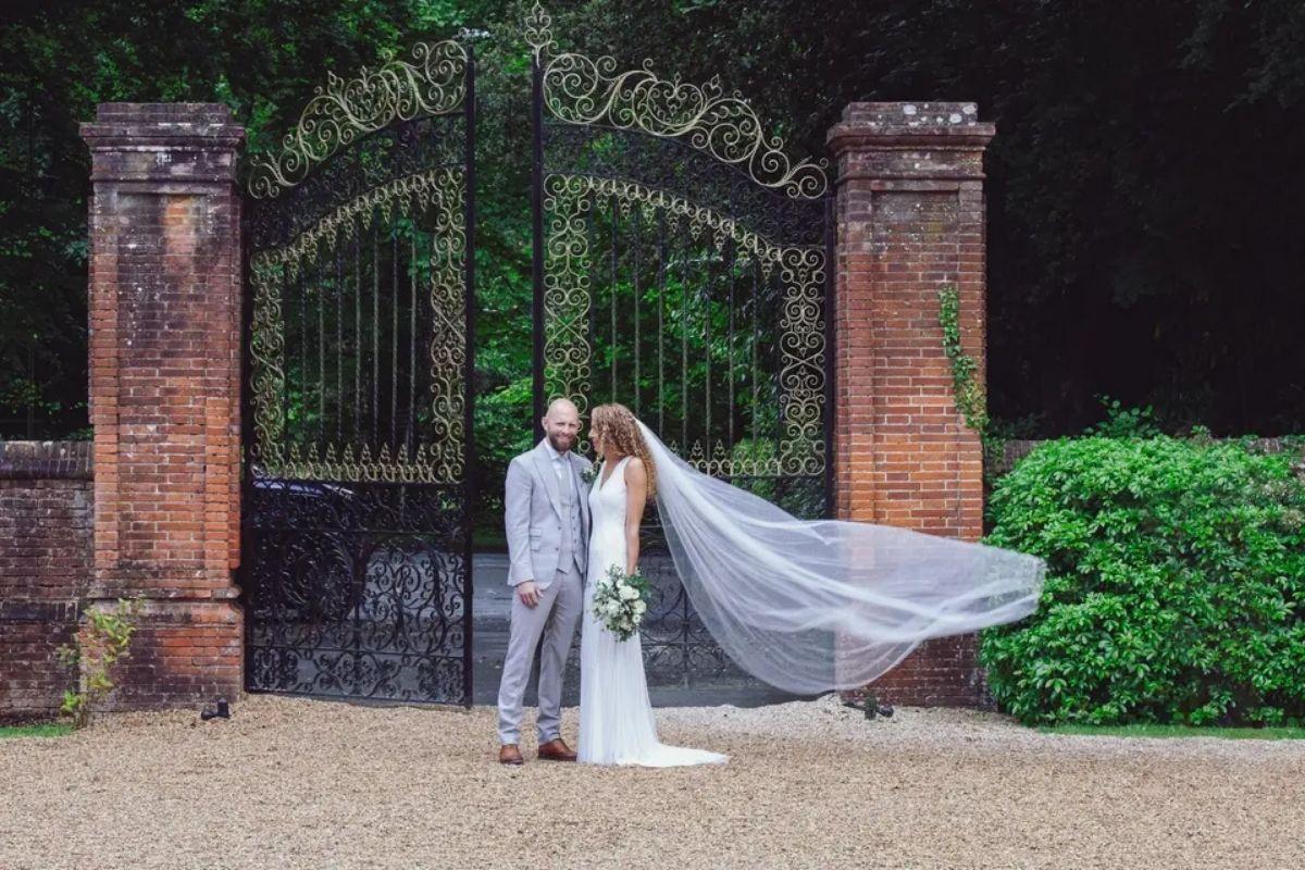 a bride poses with her veil swept away by the wind as she stands with her groom at the gates of their wedding venue