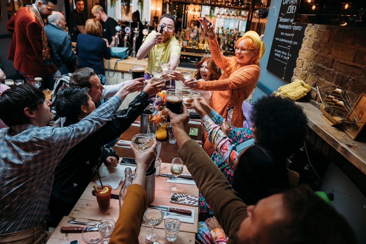 A group of friends sat on a long table in a pub toasting and cheersing