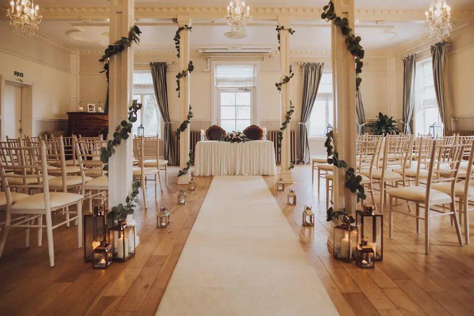 Room set for wedding ceremony with white Chiavari chairs, vine decorations, aisle lined with pillar candles, curtains and chandeliers