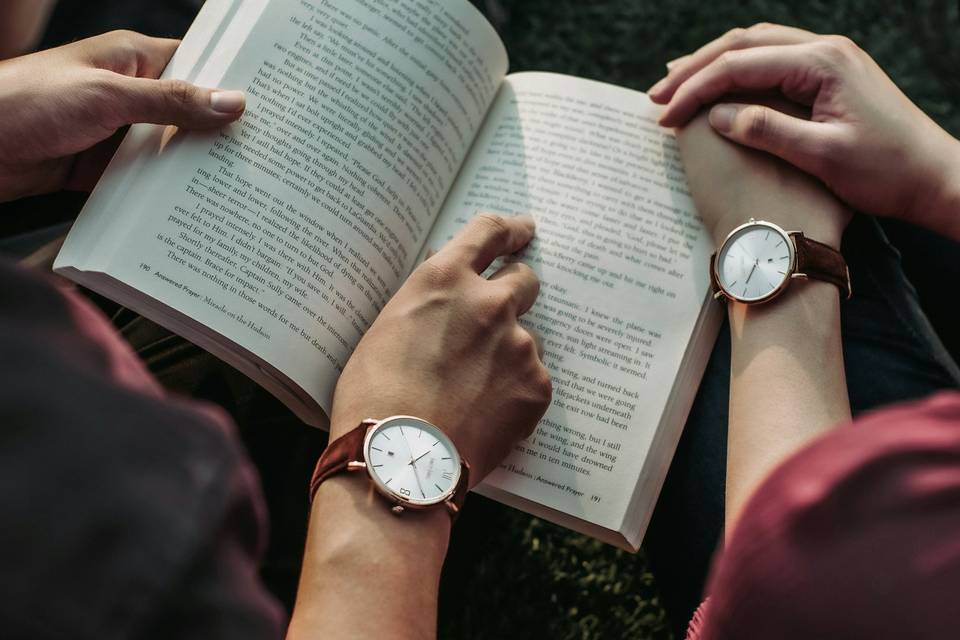 Couple wearing matching watches reading a book together on the grass, pointing out different lines to each other