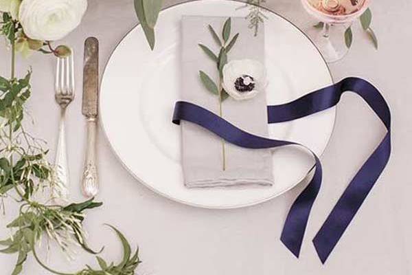 Single place setting at a table in front a lush arrangement of flowers. On the plate, there's a single grey napkin with a dark blue satin ribbon and a ranunculus flower