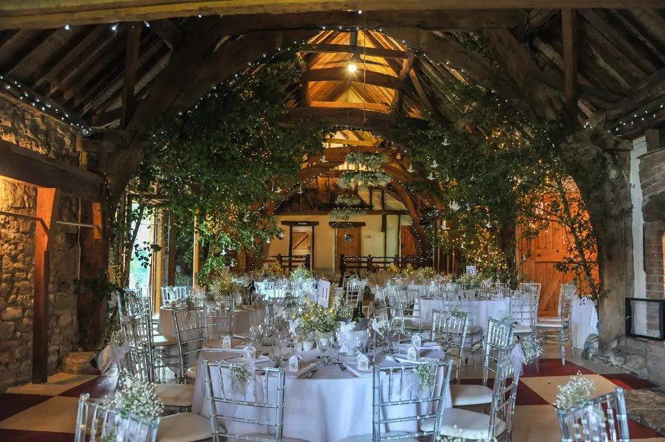 Warmly lit barn venue decorated for wedding meal with white round tables, chairs, floral white centrepieces, stone walls, exposed wooden beams and fairylights