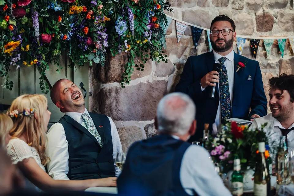 A bride and groom throw their heads back and laugh in a rustic barn wedding setting as the best man gives a speech