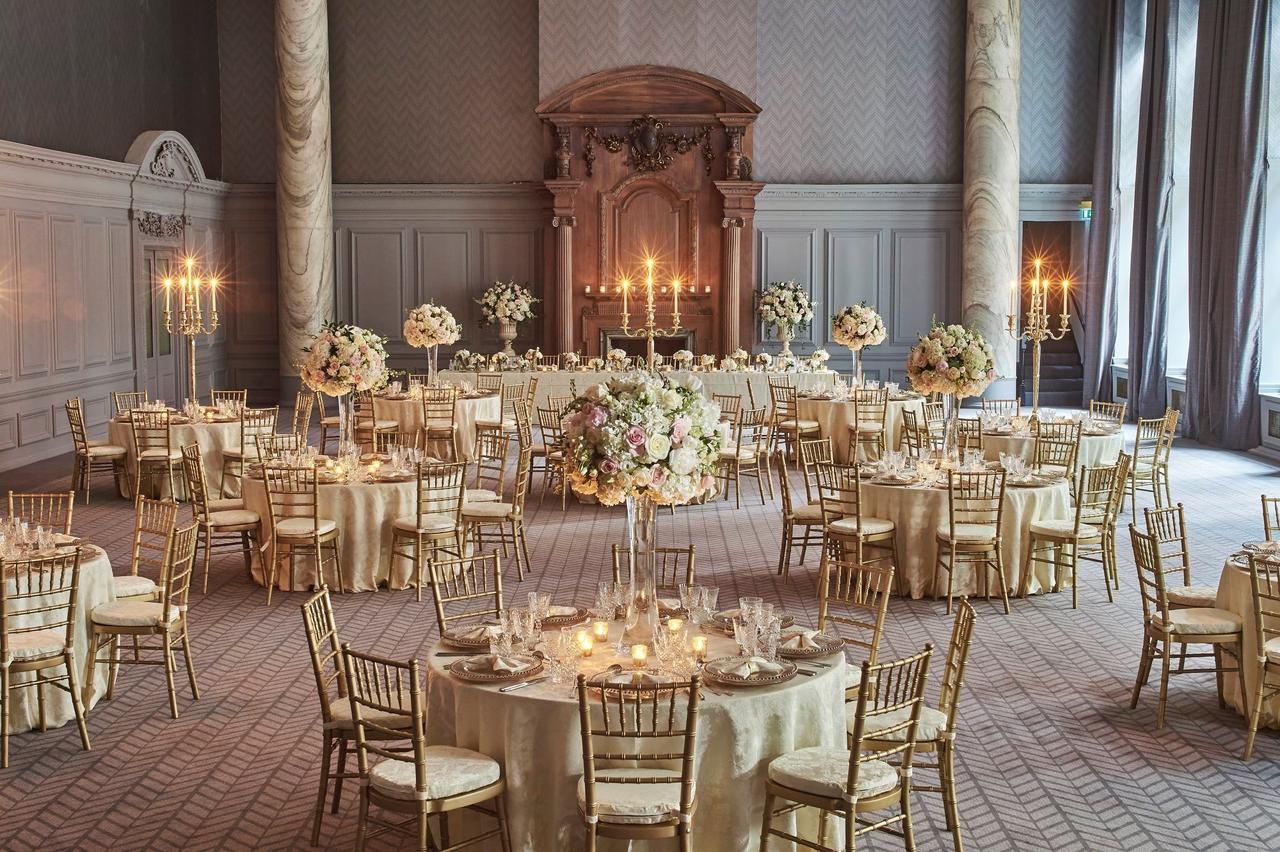 An art deco ballroom set up for a wedding breakfast with round tables, gold chiavari chairs, tall candelabra, marble pillars and a statement fireplace. 
