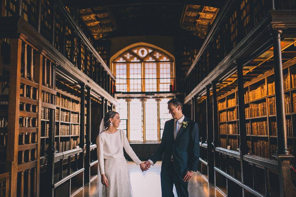 A bride and groom holding hands on their wedding day walking through a grand ancient library