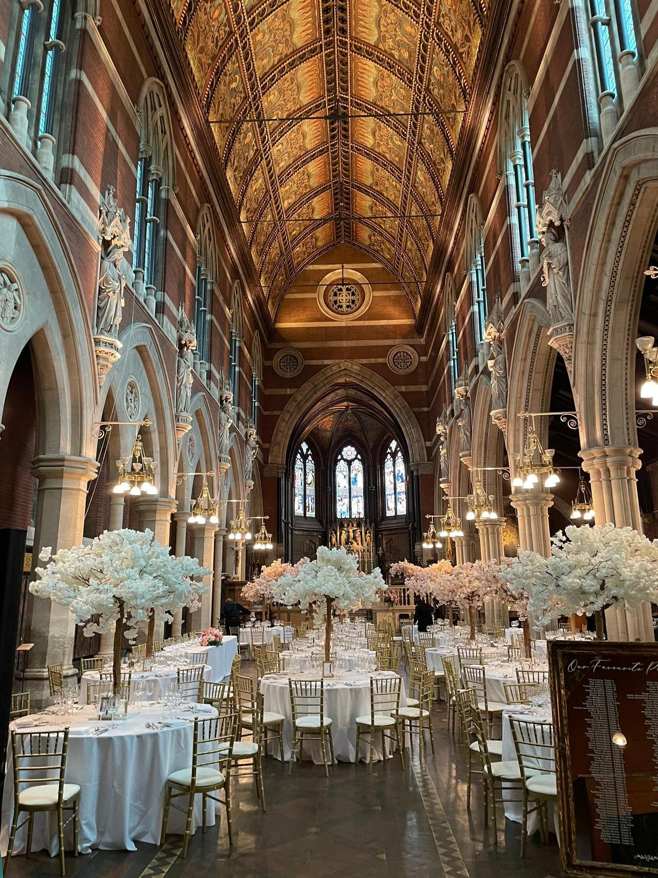Grand wedding venue with high gothic ceiling and stained glass windows, decorated with white tables, gold chairs and mini faux cherry blossom trees