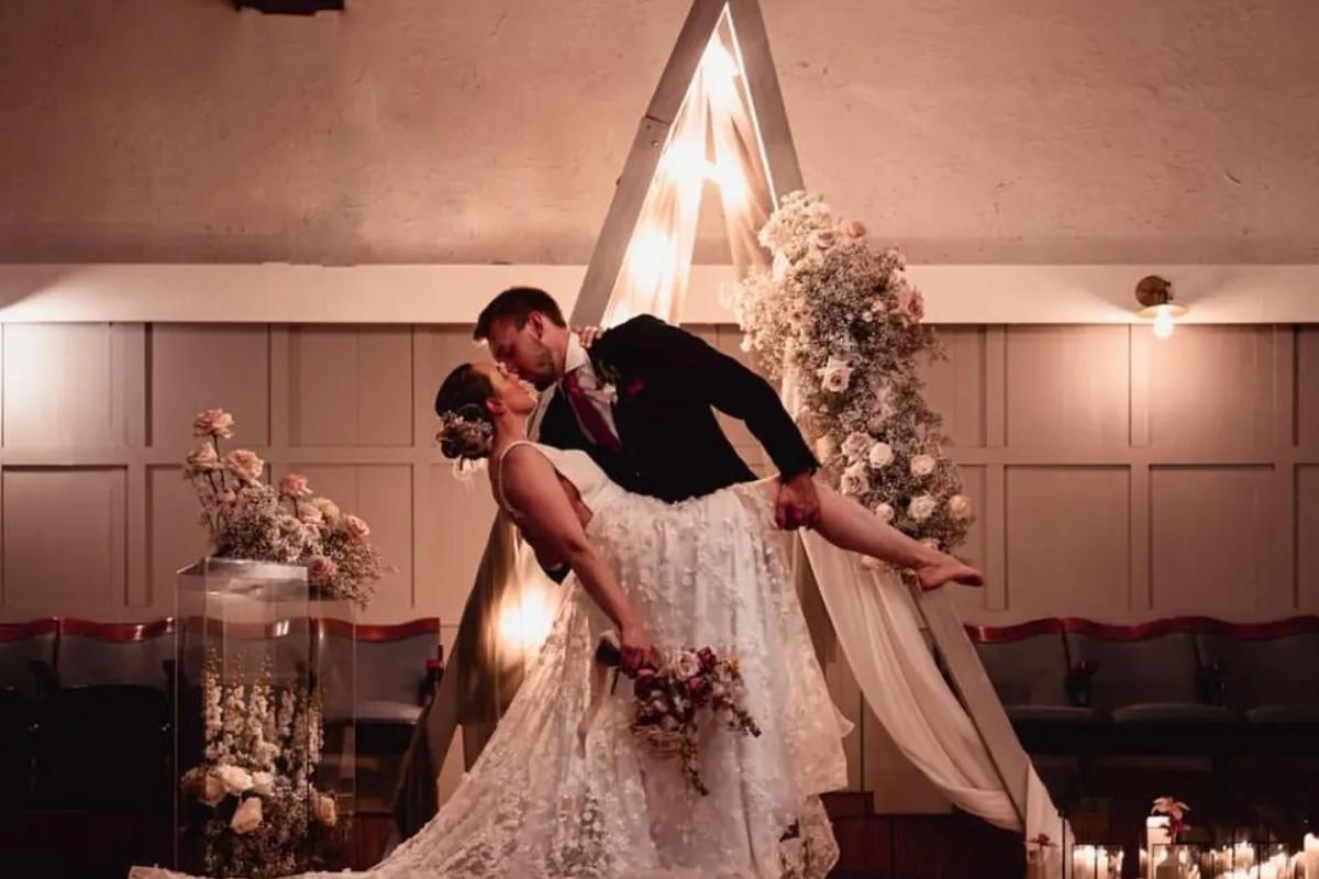 a bride leaning back while her groom holds her leg up in front of a triangular wedding arch with fairylights - zowie
