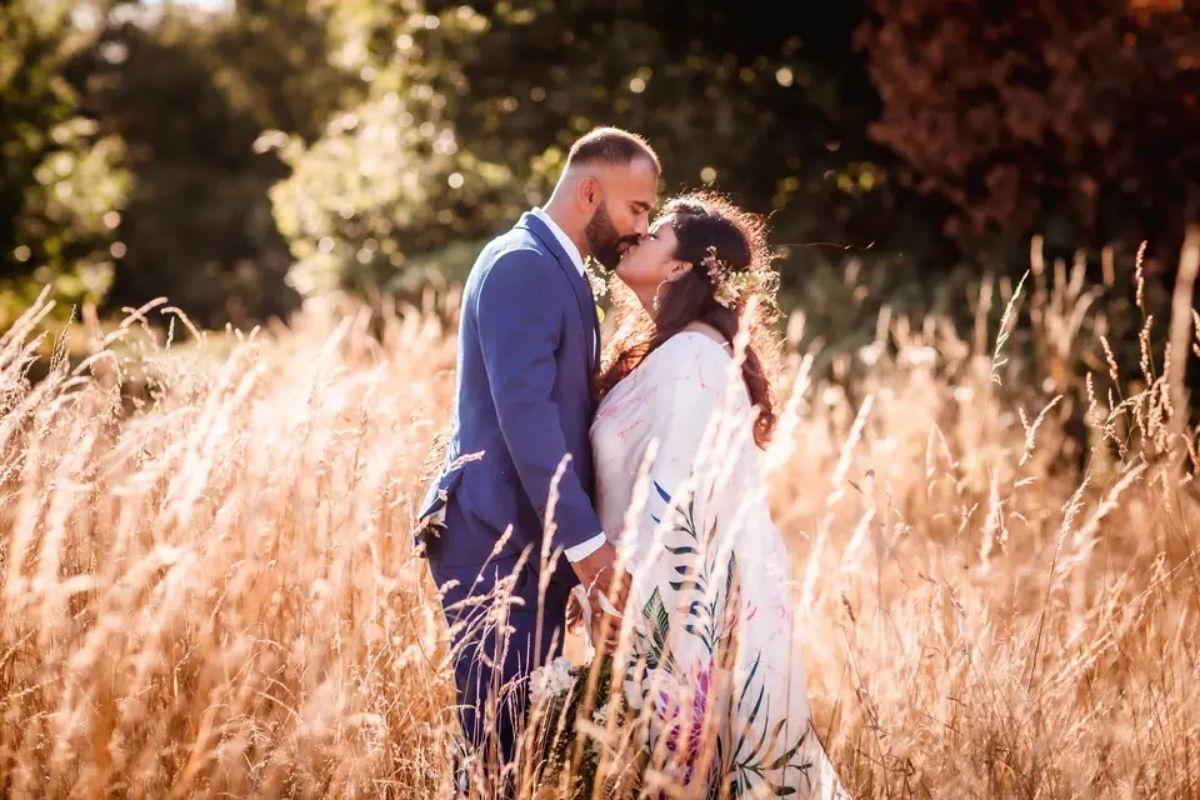 a couple in wedding attire holding hands and kissing in a field of grass with trees in the background