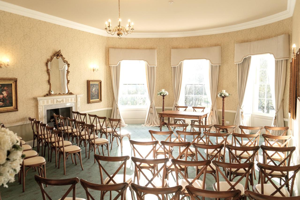 Interior of a stately home wedding venue set up for a ceremony with period fireplace, large windows and elegant white flowers.