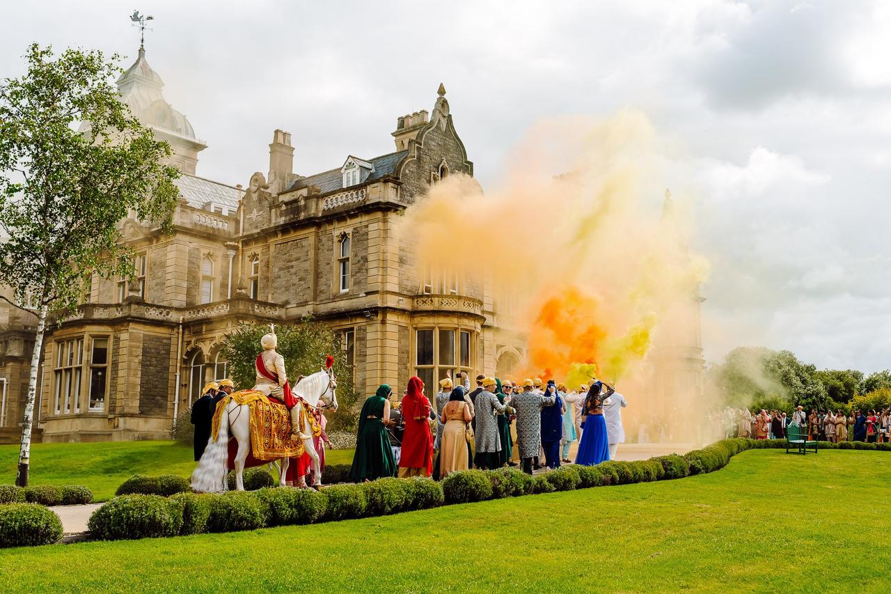 An Indian wedding, a bride and groom riding on a horse through rainbow smoke in outside stately home