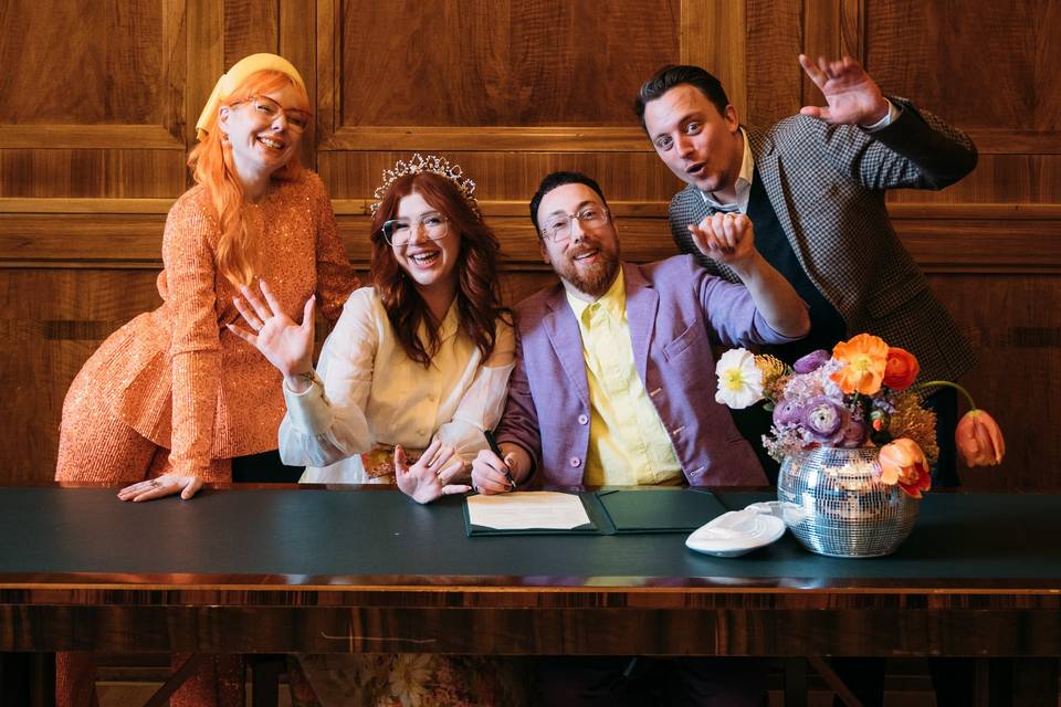 four people in colourful wedding outfits around a table in a town hall signing the register after a wedding ceremony