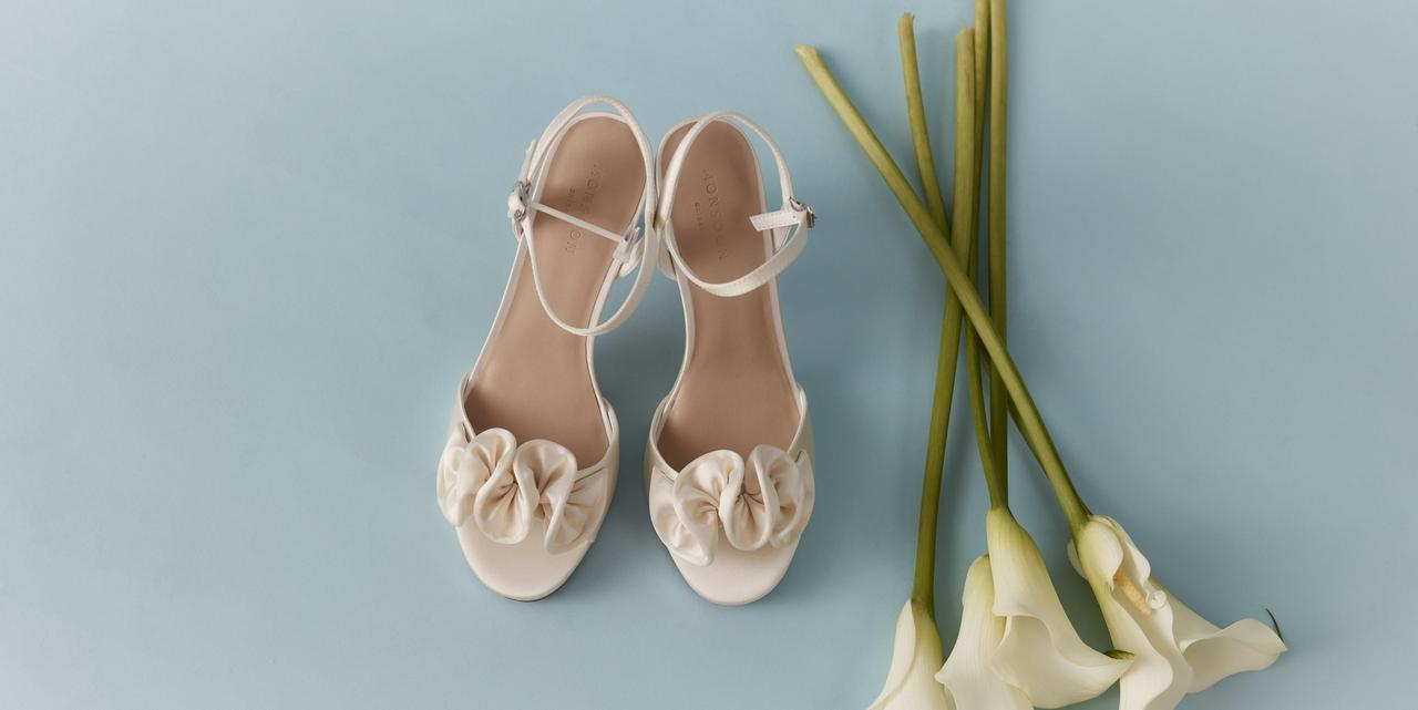 A pair of ivory platform sandals with frill detail on the front next to a bunch of white lilies on a blue background.
