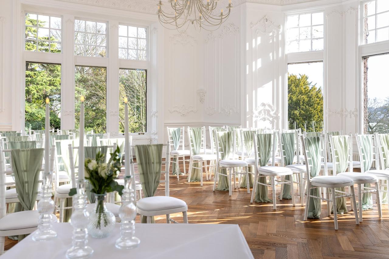 A beautiful pale blue and grey ceremony room with white chiavari chairs and large windows with views of the countryside.