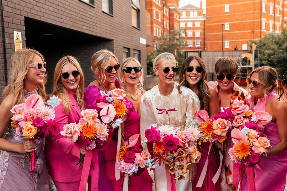 a bride surrounded by bridesmaids in pink dresses, all wearing sunglasses and holding vibrant, summery bouquets