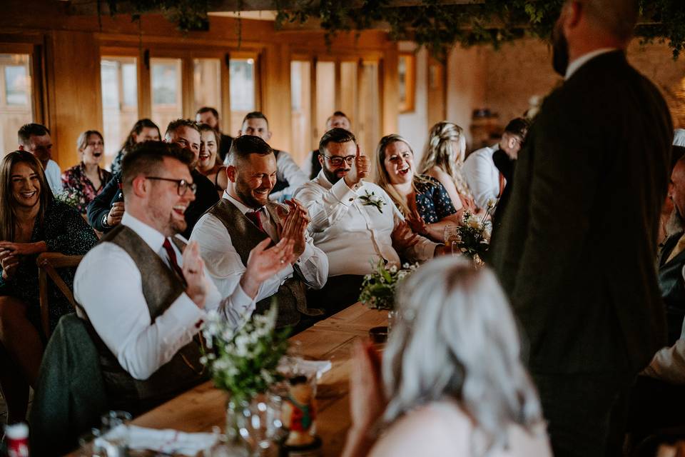 a man standing at the head of a top table making groom speech jokes in a barn wedding venue as guests laugh