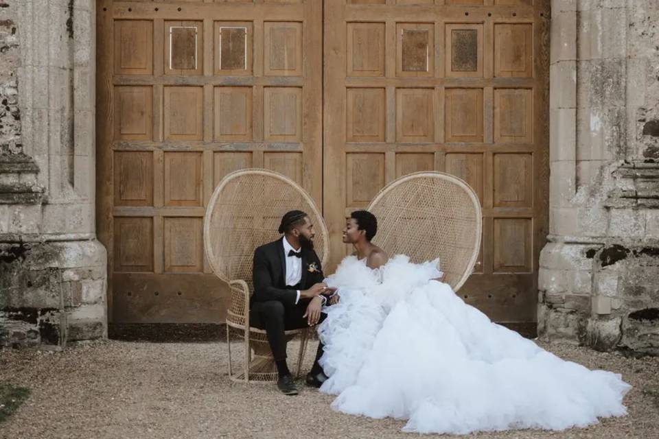 A bride and groom sitting on two wicker chairs outside a stone venue with large wooden doors