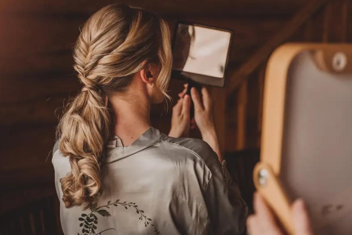A bride with blonde hair looking at her plaited low curly ponytail in the mirror as she wears a sage green dressing gown - colette