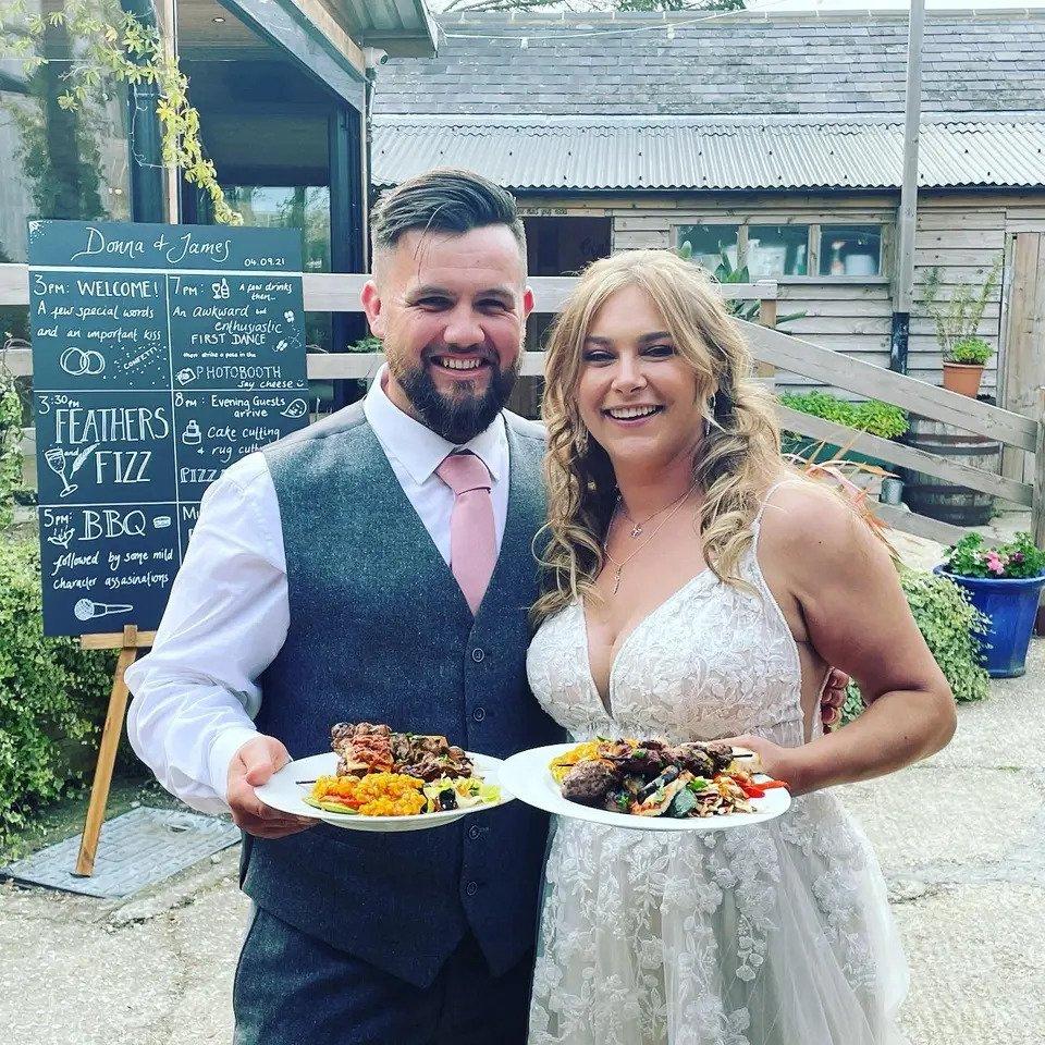 Bride and groom stand outdoors and smile with a plate of wedding food each