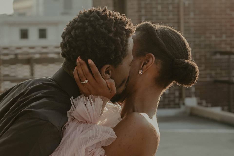 A man kisses a woman on the cheek on a roof in a city - the woman is wearing a pink chiffon off the shoulder dress