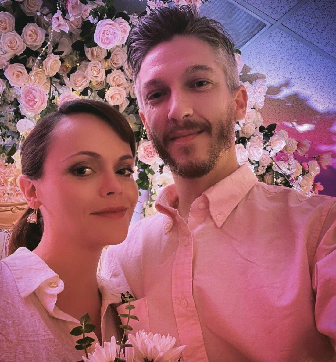 Christina Ricci in a sleek low ponytail and white collared shirt holding a white foliage bouquet next to her grey haired husband Mark Hampton wearing a white collared shirt standing in front of a pink rose flower wall