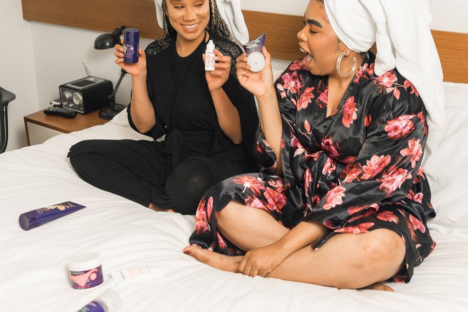 Two women sitting on a bed with their hair wrapped in towels holding skincare products about to start their skincare routine