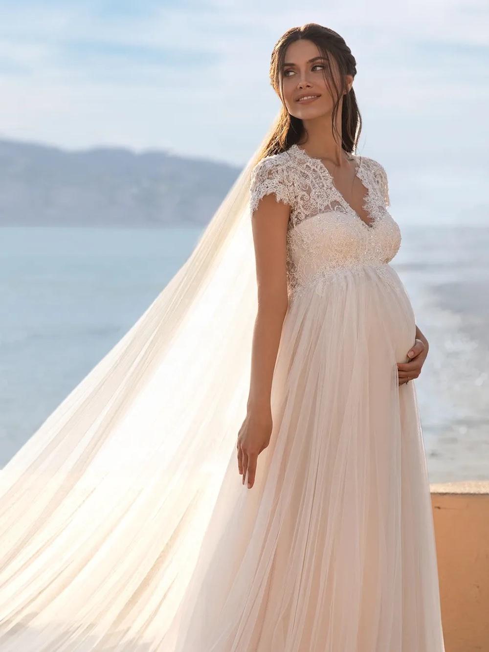 Pregnant white woman with long dark hair on a beach wearing a v necked tulle skirted white wedding dress with an embellished bodice and simple sheer white veil