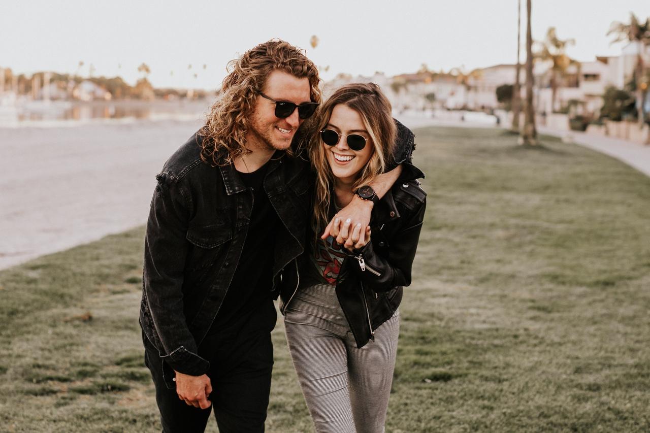 Couple in leather jackets and sunglasses laughing together. The man has his arm around the woman and she is holding his hand