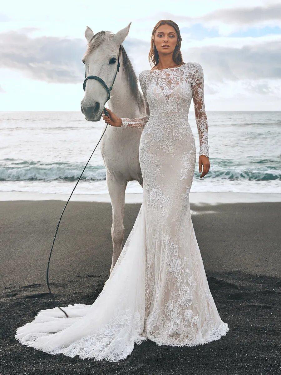 Model wearing a long sleeved lace wedding dress standing next to a horse
