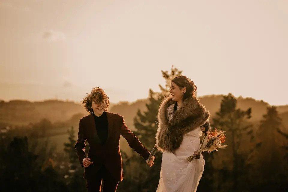 Two women hand in hand laughing together outside, one in a suit and one in a wedding dress with a faux fur wrap