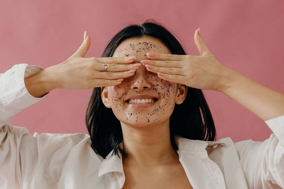Woman with a facial exfoliator applied to her skin, smiling and covering her eyes with her hands against a pink backdrop