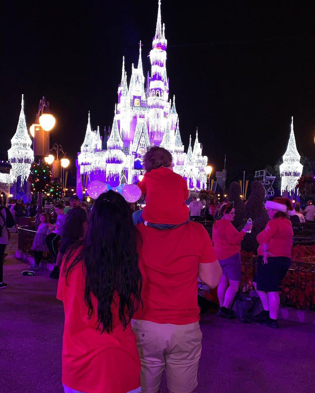 Cara Delahoyde and Nathan Masset posing at night in front of the Disney Magic Castle which is lit up. Nathan has their son Fred on his shoulders. They are all facing with their backs to the camera