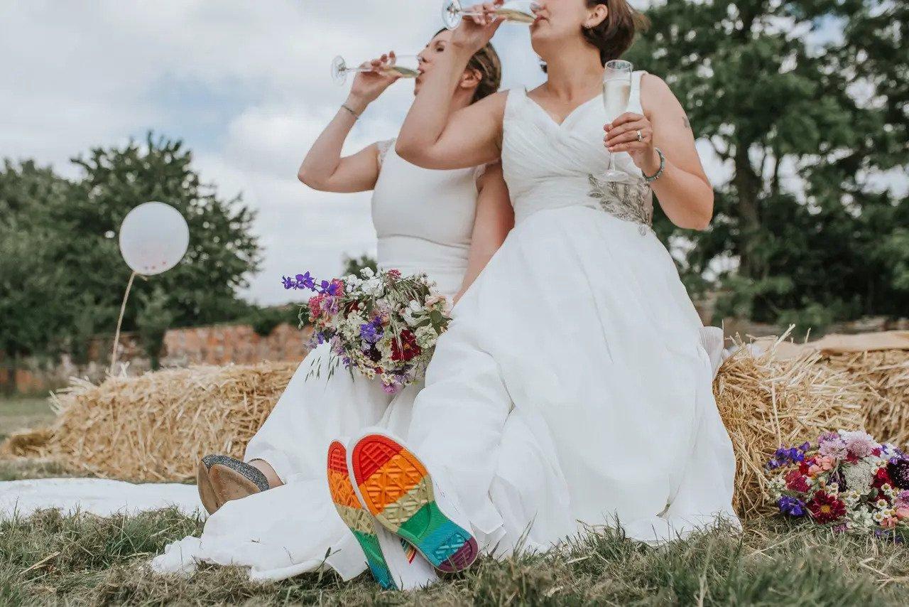 Two brides in wedding dresses sitting on a haybale drinking champagne. The rainbow soled trainers of one bride are visible peeking out from under her dress