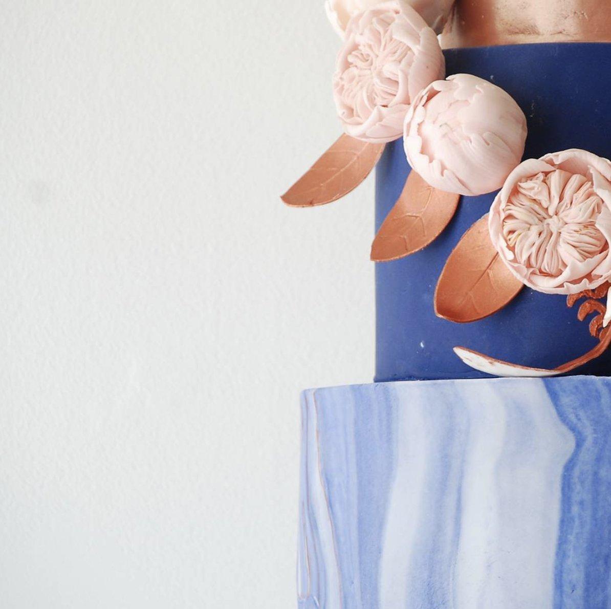 Close up of multi tiered royal blue and white marble cake with pale pink peony appliqué and copper leaves