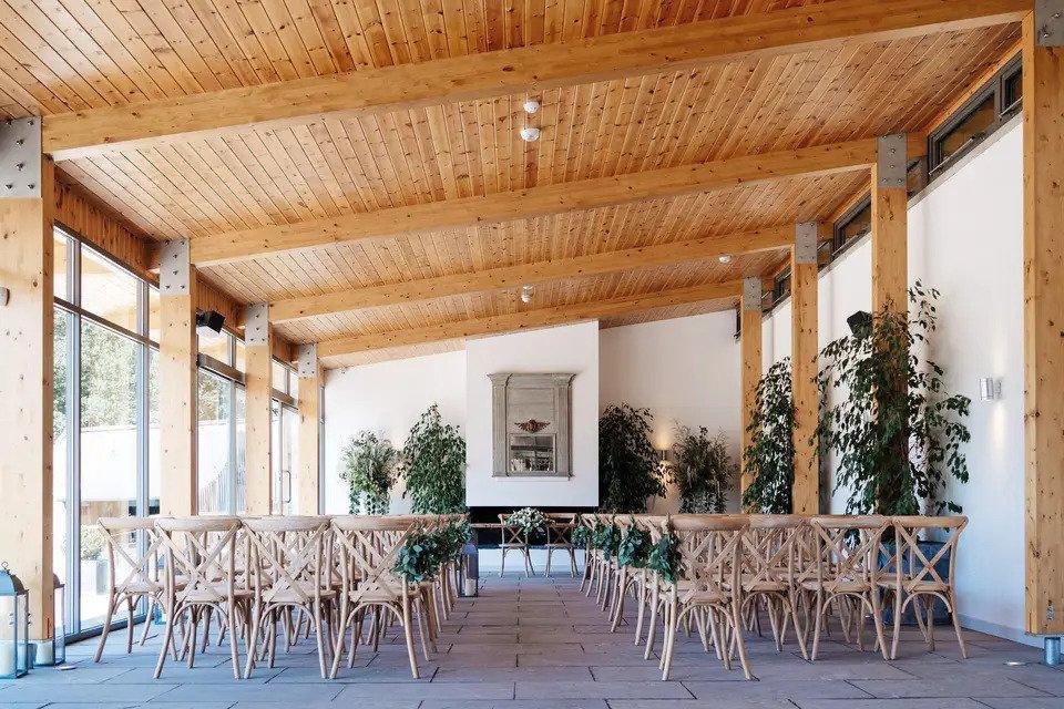 Room set for wedding ceremony with sloping wood-panelled roof, wooden beams, wooden chairs, plants and floor-to-ceiling windows